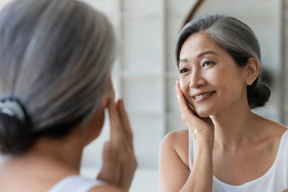 Happy 50s middle aged woman model touching face skin looking in mirror reflection.